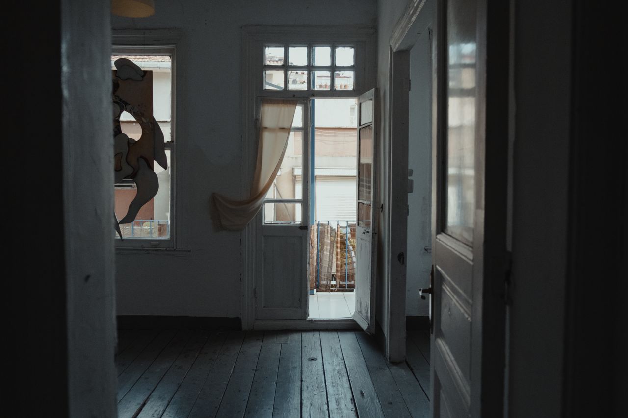 Dim interior room with wooden floorboards, tall windows, and a door standing open to a bright balcony outside.